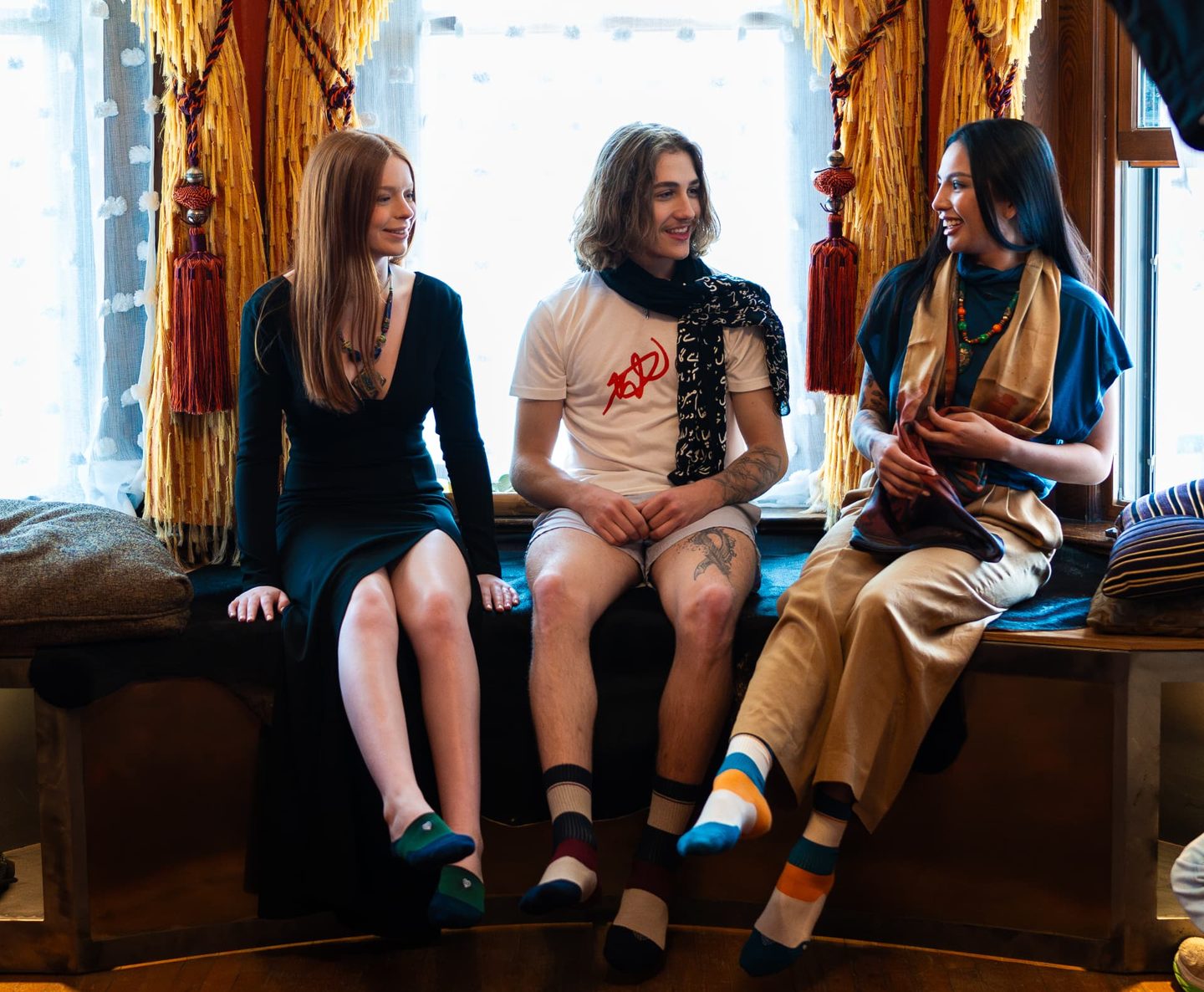 Three models seated together on a deep window bench with gold fringed curtains, laughing.